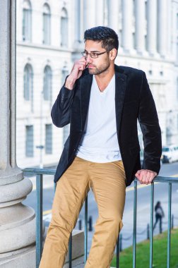 Siyah yün ceket, beyaz iç çamaşırı ve koyu sarı pantolon giyen, gözlük takan, ekmek ve bıyıklı genç bir profesyonel korkulukta oturup bir telefon konuşmasını dinliyor.