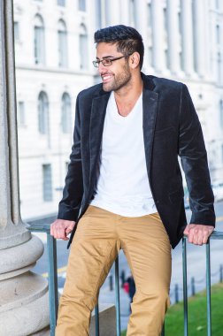 Siyah yün ceket, beyaz iç çamaşırı ve koyu sarı pantolon giyen, gözlük takan, ekmek ve bıyıklı genç bir profesyonel gülümseyerek tırabzanın kenarında oturuyor, dinleniyor ve düşünüyor.