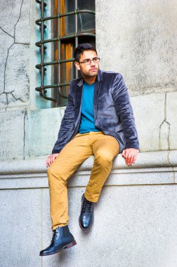 Koyu mor yün ceket, mavi kazak, koyu sarı pantolon ve deri ayakkabı giyen, gözlük takan, ekmek ve bıyıklı genç bir adam eski moda bir pencerenin yanında oturuyor, dinleniyor.