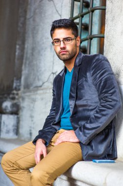 Koyu mor yün ceket, mavi kazak ve koyu sarı pantolon giyen, gözlük takan, ekmek ve bıyıklı genç bir adam dışarıda oturmuş, büyüleyici bir şekilde sana bakıyor.