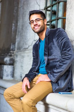 Koyu mor yün bir ceket, mavi bir süveter ve koyu sarı pantolon giyen, gözlük takan, ekmek ve bıyıklı genç bir adam dışarıda oturmuş, sana gülümsüyor.