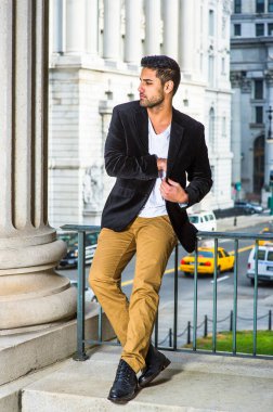 Siyah yün ceket, beyaz iç çamaşırı, koyu sarı pantolon ve deri ayakkabılar giyen, ekmek ve bıyıklı genç bir adam korkulukların yanında oturuyor, iç cebini arıyor.