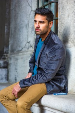 Koyu mor yün bir ceket, mavi bir süveter ve koyu sarı bir pantolon giyen, ekmek ve bıyıklı genç bir adam dışarıda oturuyor, gerçekten ileriye bakıyor.