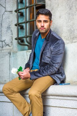 Koyu mor yün bir ceket, mavi bir süveter ve koyu sarı bir pantolon giymiş, elinde beyaz bir gül tutan, dışarıda oturan ve sana bakan bıyıklı genç bir adam.