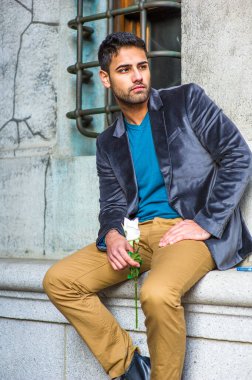 Koyu mor yün bir ceket, mavi bir süveter ve koyu sarı bir pantolon giymiş, elinde beyaz bir gül tutan, dışarıda oturan ve sana bakan bıyıklı genç bir adam.