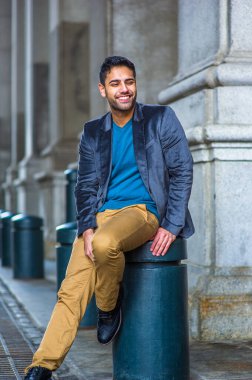 Mavi yün ceket, kazak ve koyu sarı pantolon giyen, ekmek ve bıyıklı genç bir adam eski moda bir sokakta oturuyor, gülümsüyor.