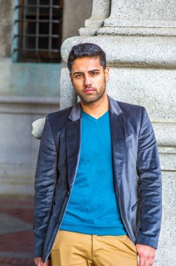 Koyu mor yün ceket ve mavi kazak giyen, ekmek ve bıyıklı genç yakışıklı bir adam bir sütunun yanında durup düşünceli bir şekilde sana bakıyor.