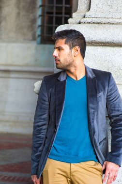 Koyu mor yün ceket ve mavi kazak giyen, ekmek ve bıyıklı genç yakışıklı bir adam bir sütunun yanında duruyor, gözlerini kısıyor ve etrafına bakıyor.