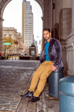 Yünlü ceket, süveter, koyu sarı pantolon ve deri ayakkabı giyen, gözlük takan, dışarıda oturan, bıyıklı ve ekmeği olan genç bir profesyonel,