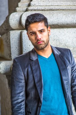 Koyu mor yün ceket ve mavi kazak giyen, ekmek ve bıyıklı genç yakışıklı bir adam bir sütunun yanında durup düşünceli bir şekilde sana bakıyor.