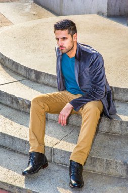 Koyu mor yün ceket, koyu sarı pantolon ve deri çizme ayakkabı giyen, ekmek ve bıyıklı genç bir Orta Doğulu, daire basamaklarında oturuyor, rahatlıyor ve düşünüyor.