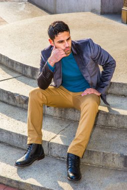Koyu mor yün ceket, koyu sarı pantolon ve deri çizme ayakkabı giyen, ekmek ve bıyıklı genç bir Orta Doğulu, daire basamaklarında oturuyor, rahatlıyor ve düşünüyor.