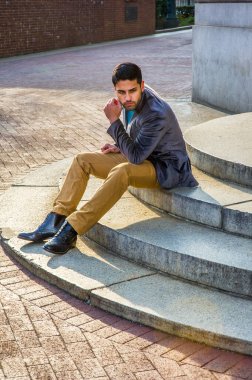 Koyu mor yün ceket, koyu sarı pantolon ve deri çizme ayakkabı giyen, ekmek ve bıyıklı genç bir Orta Doğulu, daire basamaklarında oturuyor, rahatlıyor ve düşünüyor.