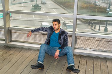 Cam bir duvara yaslanmış, sakallı ve bıyıklı genç bir adam yere oturmuş derin derin düşünüyor. Arka plan bir caddenin üzerinde.