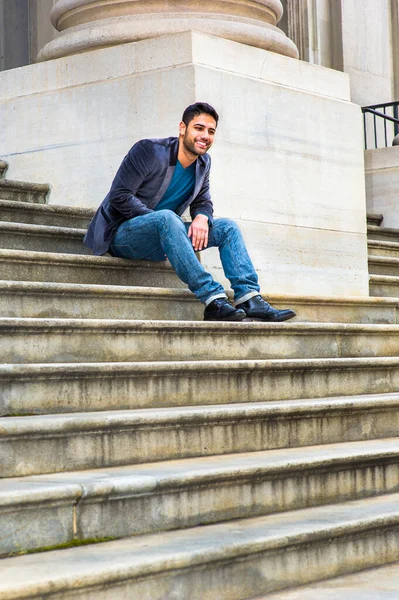Koyu mor yün ceket, mavi kot pantolon ve siyah deri ayakkabılar giyen, ekmek ve bıyıklı genç bir Orta Doğulu, basamaklarda oturuyor, düşünüyor ve gülüyor..