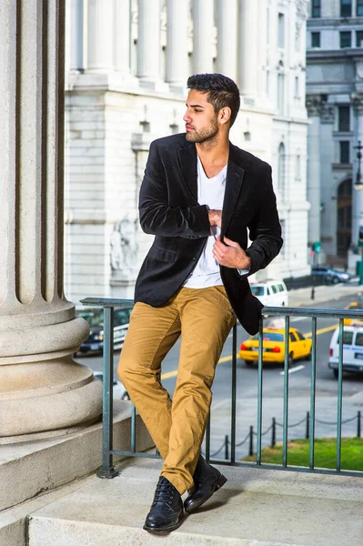 Siyah yün ceket, beyaz iç çamaşırı, koyu sarı pantolon ve deri ayakkabılar giyen, ekmek ve bıyıklı genç bir adam korkulukların yanında oturuyor, iç cebini arıyor.