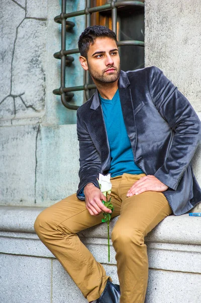 Koyu mor yün bir ceket, mavi bir süveter ve koyu sarı bir pantolon giymiş, elinde beyaz bir gül tutan, dışarıda oturan ve sana bakan bıyıklı genç bir adam.