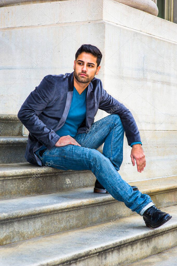 Dressing in a dark purple woolen blazer,  blue jeans and black leather shoes, a young handsome Middle Eastern guy with bread and mustache is sitting on steps outside an office, thoughtfully looking around