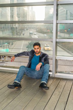 Cam bir duvara karşı, sakallı ve bıyıklı genç bir adam yerde oturuyor. Düşünceli bir şekilde sana bakıyorum. Arka plan kalabalık bir sokak sahnesi.