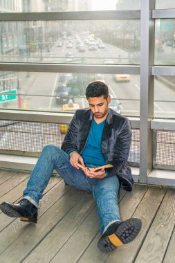 Cam bir duvara yaslanmış, sakallı ve bıyıklı genç bir adam yerde oturmuş kitap okuyor. Arka plan kalabalık bir sokak sahnesi.