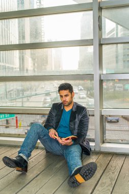 Cam bir duvara yaslanmış, sakallı ve bıyıklı genç bir adam yerde oturmuş kitap okuyor. Arka plan kalabalık bir sokak sahnesi.