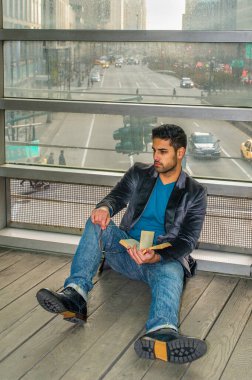 Cam bir duvara yaslanmış, elinde gözlük olan genç bir öğrenci yerde oturmuş kitap okuyor ve düşünüyor. Arka plan kalabalık bir sokak sahnesi.