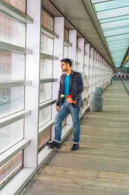 Gözlük takıyor, kırmızı bir kitap ve beyaz bir çiçek tutuyor, sakallı ve bıyıklı genç bir adam yürüyüş yolunda ve cam duvara bakıyor, ciddi ciddi dışarı bakıyor..