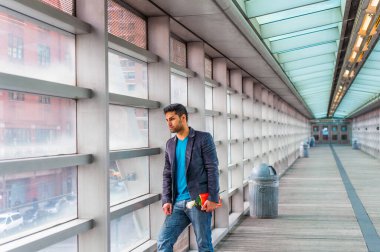 Elinde kırmızı bir kitap ve beyaz bir çiçek, sakallı ve bıyıklı genç bir adam cam duvara doğru yürüyor, ciddi ciddi dışarı bakıyor.