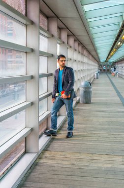 Gözlük takıyor, kırmızı bir kitap ve beyaz bir çiçek tutuyor, sakallı ve bıyıklı genç bir adam yürüyüş yolunda duruyor, cam duvara yaslanmış,