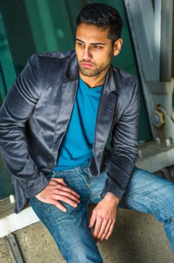 Koyu mor yün bir ceket, mavi bir kazak ve kot pantolon giymiş sakallı ve bıyıklı genç yakışıklı bir adam bir yapının yanında oturuyor ve derin bir şekilde düşünüyor.