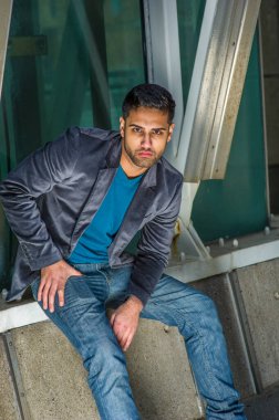 Koyu mor yün bir ceket, mavi bir kazak ve kot pantolon giymiş, sakallı ve bıyıklı genç bir adam köşede oturuyor, eğiliyor, düşünceli bir şekilde sana bakıyor.