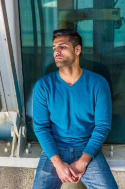 Mavi bir kazak ve kot pantolon giymiş sakallı ve bıyıklı genç yakışıklı bir adam köşede oturuyor, düşüncelere dalmış.