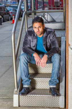 Koyu mor yün ceket, kot pantolon ve deri ayakkabılar giyen, sakallı ve bıyıklı yakışıklı genç bir işçi bir ofis binasının merdivenlerinde oturuyor, eğiliyor, dinleniyor.