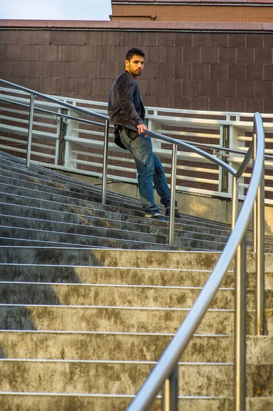 身穿深紫色羊毛衫和牛仔裤的小伙子 留着胡子 站在楼梯上的铁栏杆上 在夕阳西下 望着你 — 图库照片