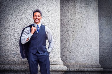 Açık gri bir gömlek, koyu mavi bir yelek, bir kravat ve omuzundan çıkarılan ceketle, yakışıklı, orta yaşlı bir iş adamı dışarıda kolonların yanında duruyor, gülümsüyor ve sana bakıyor.