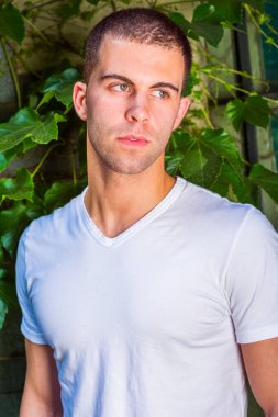 Beyaz V yaka tişört giyen, kısa saçlı genç bir adamın portresi. Arka plan yeşil sarmaşık yaprakları. Kapat.