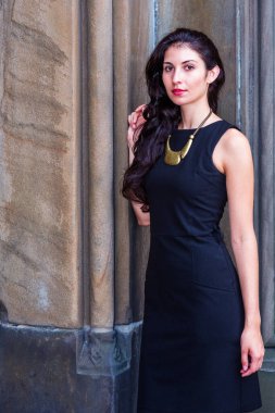 Güzel Genç Bir Kadının Portresi. Kolsuz siyah bir elbise, altın kolyeli bir kolye, genç ve seksi bir iş kadını duvarın önünde durup düşünüyor, mola veriyor.