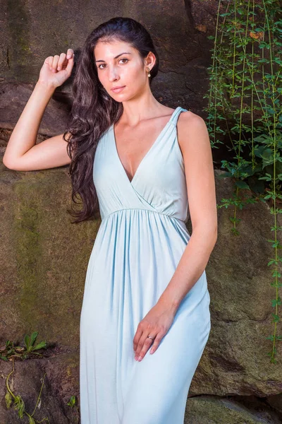 Portrait of Pretty Lady. Dressing in a light blue long dress, a young sexy woman with long curly hair is standing by a rocky wall with long green leaves, charmingly looking at you in hot summer