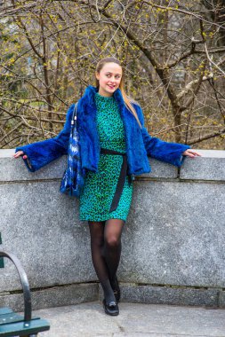 Dressing in blue faux fur coat, patterned dress, carrying a bag, a pretty girl is standing by a fence, smiling. Small yellow buds on branches of trees in the background
