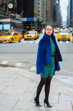 Dressing in a blue faux fur coat,  patterned dress, black leggings and leather shoes, a pretty woman is standing on a busy street of a big city, looking around