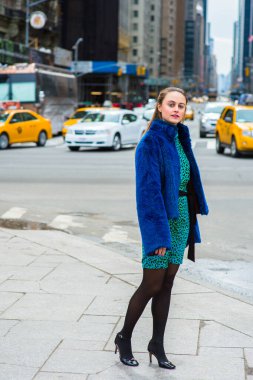 Dressing in a blue faux fur coat,  patterned dress, black leggings and leather shoes, a pretty woman is standing on a busy street of a big city, looking around