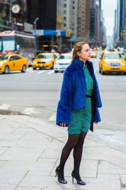 Dressing in a blue faux fur coat,  patterned dress, black leggings and leather shoes, a pretty woman is standing on a busy street of a big city, looking around