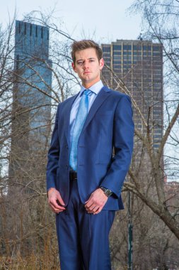 Dressing formally a young professional is standing outside and thoughtfully looking at you. The background is two high business buildings