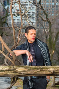 Dressing in a black shirt, tie and long scarf, a young handsome guy is pondering at small woods  in winter.