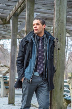 Dressing in a pea coat, woolen jacket, shirt, scarf, tie, a young handsome student is standing by a pillar of woolen structure in winter, looking forward and waiting for you