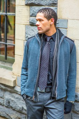 Dressing in woolen jacket with shoulder pads, tie, long scarf and gloves, a young handsome guy is standing against the wall, looking forward