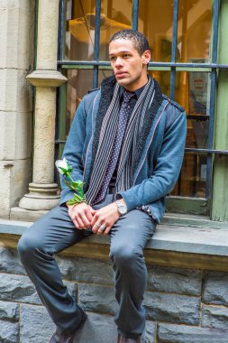 Dressing in woolen jacket with shoulder pads, tie, long scarf, holding a white flower, a young handsome guy with short, curly hair is sitting on the frame of a window, waiting for you