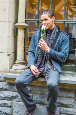 Dressing in woolen jacket with shoulder pads, tie, long scarf, holding a white flower, a young handsome guy with short, curly hair is sitting on the frame of a window, smelling and thinking.