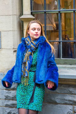 Dressing in a blue faux fur plus size jacket, patterned dress and scarf, a pretty girl is standing by a window in winter, looking forward and waiting for you
