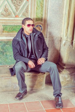 Young man wearing sunglass, sitting against wall in winter, thinking, wearing in woolen jacket, tie, long scarf, black pants, leather shoes